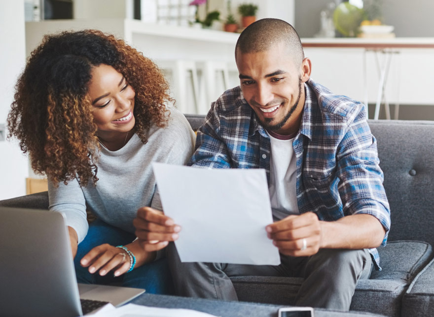 Female with computer and male with paper in hand, smiling, in living room.